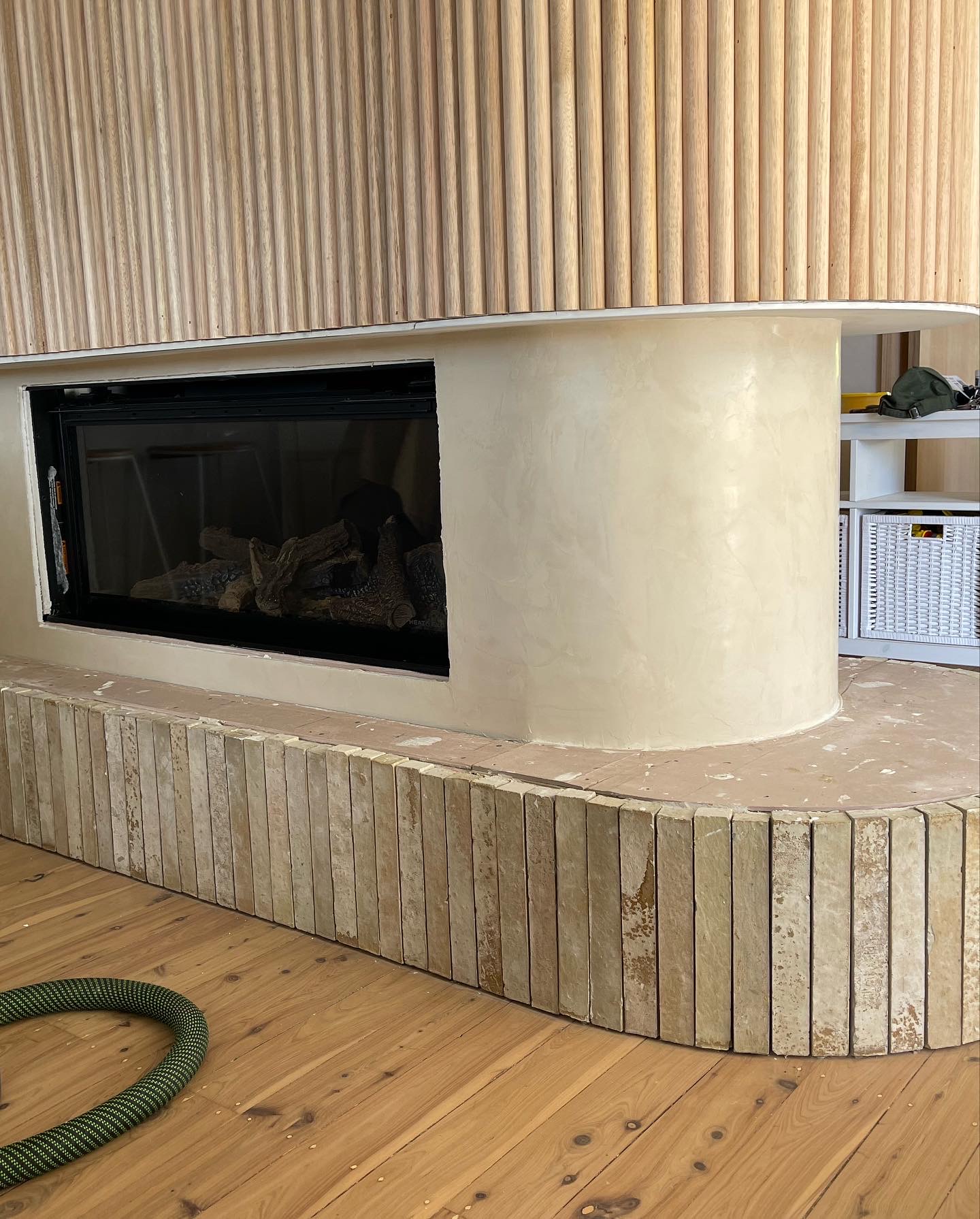 Curved beige plaster fireplace surround with a black-framed firebox; vertical wooden slat wall above and a coiled green hose on a wooden floor nearby.