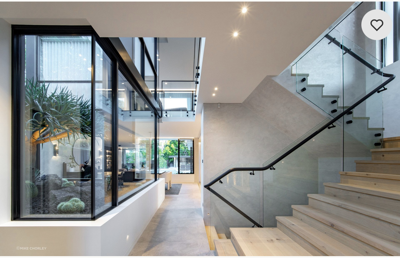 Modern indoor lobby with a glass-walled atrium, visible courtyard plants, and a wooden staircase with glass railing on the right.